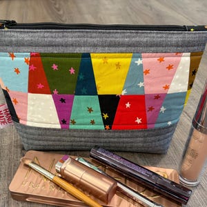 Pu&ograve; includere: Un beauty case fatto a mano con un design patchwork con vari colori e motivi a stella. La borsa ha una cerniera grigia e un tirazip rosso. I prodotti per il trucco, tra cui un flacone di fondotinta, una palette di ombretti e mascara, sono accanto alla borsa.