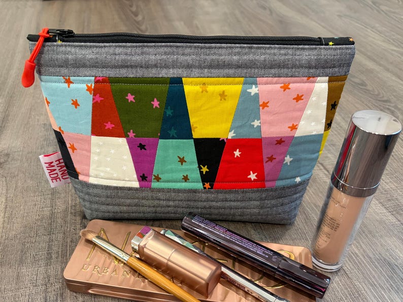 Pu&ograve; includere: Un beauty case fatto a mano con un design patchwork con vari colori e motivi a stella. La borsa ha una cerniera grigia e un tirazip rosso. I prodotti per il trucco, tra cui un flacone di fondotinta, una palette di ombretti e mascara, sono accanto alla borsa.