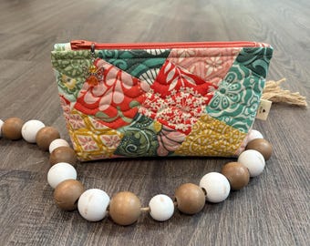 May include: A quilted fabric pouch featuring a patchwork design of floral and geometric patterns in shades of red, green, yellow, and pink. The pouch has an orange zipper with a small angel charm and a beige tassel, accompanied by a wooden bead garland.
