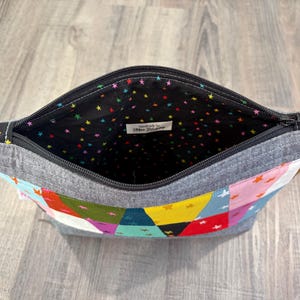 Pu&ograve; includere: Pochette con cerniera aperta con un design patchwork. L'esterno presenta un mix di forme geometriche colorate e stelle. L'interno &egrave; nero con stelle colorate. La cerniera ha un tirante rosso.