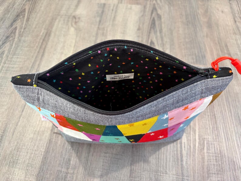 Pu&ograve; includere: Pochette con cerniera aperta con un design patchwork. L'esterno presenta un mix di forme geometriche colorate e stelle. L'interno &egrave; nero con stelle colorate. La cerniera ha un tirante rosso.