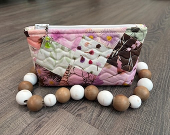 May include: A quilted fabric pouch featuring a patchwork of floral patterns in shades of pink, brown, and white. The pouch has a white zipper closure with a small green angel charm attached and is surrounded by a string of alternating wooden beads.