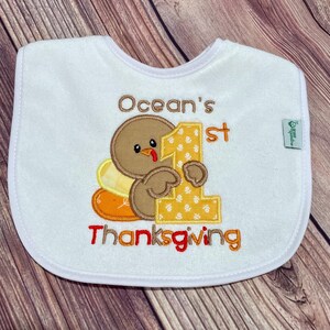 Puede incluir: Un babero blanco para beb&eacute; con un pavo marr&oacute;n bordado que sostiene un n&uacute;mero uno grande de color amarillo. El texto dice Ocean's 1st Thanksgiving en bordado marr&oacute;n y naranja. Una peque&ntilde;a etiqueta verde muestra la marca Green Sprouts.