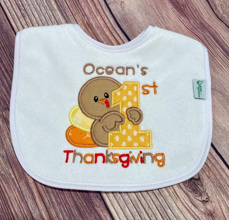 Puede incluir: Un babero blanco para beb&eacute; con un pavo marr&oacute;n bordado que sostiene un n&uacute;mero uno grande de color amarillo. El texto dice Ocean's 1st Thanksgiving en bordado marr&oacute;n y naranja. Una peque&ntilde;a etiqueta verde muestra la marca Green Sprouts.