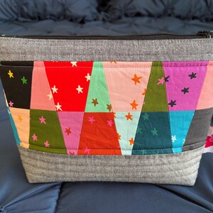 Pu&ograve; includere: Un astuccio rettangolare grigio con cerniera e design patchwork. Il patchwork presenta triangoli colorati con accenti di stelle bianche. L'astuccio ha una base e una parte superiore grigie e una piccola etichetta laterale.