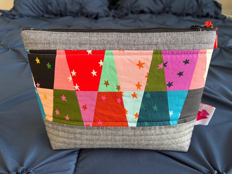 Pu&ograve; includere: Un astuccio rettangolare grigio con cerniera e design patchwork. Il patchwork presenta triangoli colorati con accenti di stelle bianche. L'astuccio ha una base e una parte superiore grigie e una piccola etichetta laterale.