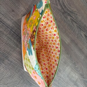 K&ouml;nnte beinhalten: Ein Rei&szlig;verschlusst&auml;schchen mit einer Patchwork-Au&szlig;enseite aus verschiedenen Blumen- und geometrischen Mustern in Gelb-, Orange-, Blau-, Gr&uuml;n- und Rosat&ouml;nen. Das Innenfutter besteht aus einem hellcremefarbenen Stoff mit einem Muster aus kleinen rosa und gelben Bl&uuml;ten.