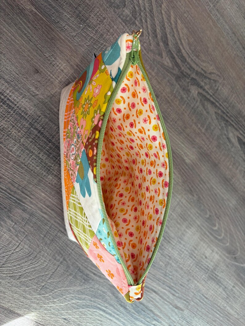 K&ouml;nnte beinhalten: Ein Rei&szlig;verschlusst&auml;schchen mit einer Patchwork-Au&szlig;enseite aus verschiedenen Blumen- und geometrischen Mustern in Gelb-, Orange-, Blau-, Gr&uuml;n- und Rosat&ouml;nen. Das Innenfutter besteht aus einem hellcremefarbenen Stoff mit einem Muster aus kleinen rosa und gelben Bl&uuml;ten.