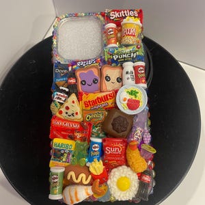 May include: A colorful phone case decorated with miniature food and candy items, including Skittles, popcorn, Sour Punch, Starburst, Cheetos, 100 Grand, Haribo, Pringles, Sun Chips, Coca-Cola, and more.