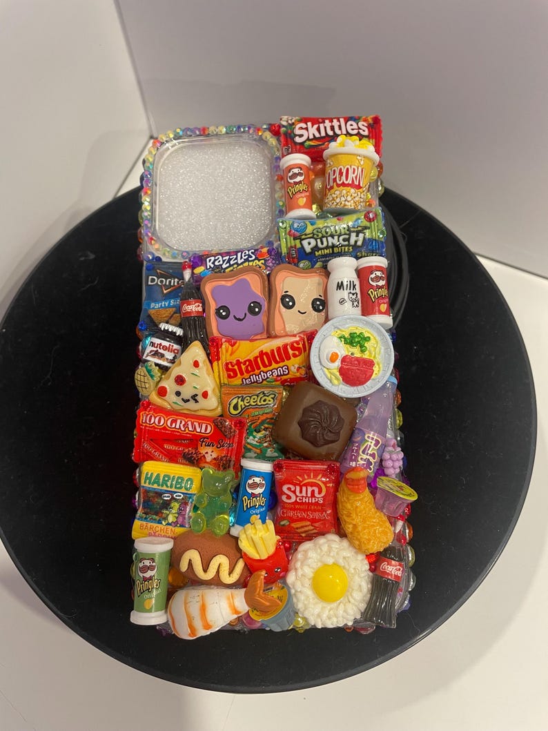 May include: A colorful phone case decorated with miniature food and candy items, including Skittles, popcorn, Sour Punch, Starburst, Cheetos, 100 Grand, Haribo, Pringles, Sun Chips, Coca-Cola, and more.