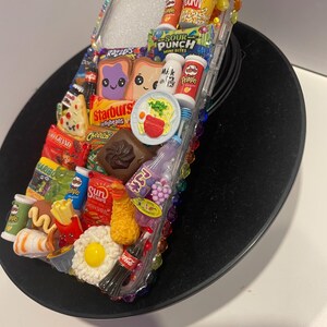May include: A clear phone case decorated with miniature food and candy items, including a bag of Skittles, a box of popcorn, a bag of Sour Punch, a box of Pringles, a bag of Cheecos, a box of Starburst jellybeans, a bag of Haribo gummy candies, a can of Coca-Cola, a bag of Sun Chips, a box of milk, a bag of Doritos, a bag of 3D Grand, a box of pizza, a bag of Razzles, a box of eggs, a bag of fries, a hot dog, a sushi roll, a fried egg, a bowl of ramen, and two slices of bread with a purple and a yellow cartoon face.