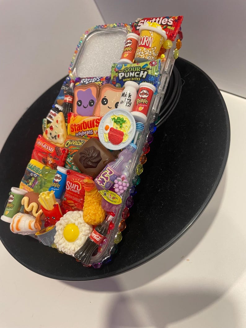 May include: A clear phone case decorated with miniature food and candy items, including a bag of Skittles, a box of popcorn, a bag of Sour Punch, a box of Pringles, a bag of Cheecos, a box of Starburst jellybeans, a bag of Haribo gummy candies, a can of Coca-Cola, a bag of Sun Chips, a box of milk, a bag of Doritos, a bag of 3D Grand, a box of pizza, a bag of Razzles, a box of eggs, a bag of fries, a hot dog, a sushi roll, a fried egg, a bowl of ramen, and two slices of bread with a purple and a yellow cartoon face.