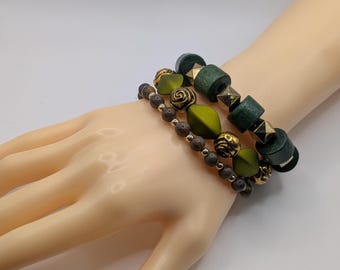 Può includere: Tre braccialetti con una varietà di perline e forme. Un braccialetto presenta perline di legno verde scuro con dettagli dorati. Un altro ha perline verde oliva e oro. Il terzo braccialetto ha piccole perline grigio scuro.