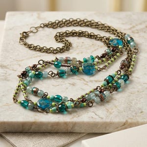 Puede incluir: Collar de varias hebras con cuentas turquesas, verde azulado y marrones. El collar tiene una cadena y un cierre de color bronce. Las cuentas son de varias formas y tamaños, algunas con un aspecto texturizado. El collar se exhibe sobre una superficie de mármol.