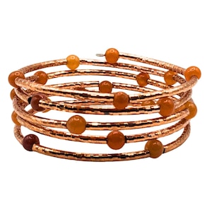 May include: A spiral bracelet with copper-colored metal bands and orange-brown beads. The bracelet wraps around multiple times, creating a layered effect. The beads are evenly spaced along the bands, adding a touch of color and texture to the design.