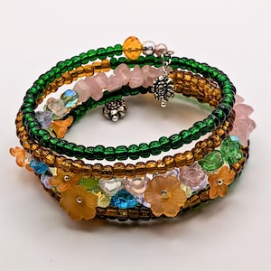 May include: A multi-strand wrap bracelet featuring green and amber glass seed beads, interspersed with translucent flower-shaped beads in orange, pink, and blue, raw pink stone chips, and two small silver-toned metal flower charms.