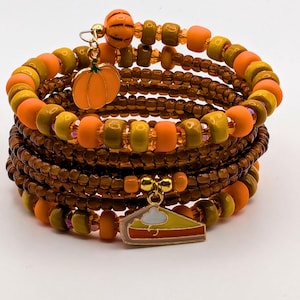 Könnte beinhalten: Ein Wickelarmband mit braunen, orangefarbenen und gelben Perlen. Das Armband hat einen Kürbis-Anhänger und einen Kuchenstück-Anhänger. Die Perlen sind in mehreren Strängen angeordnet, wodurch ein geschichteter Effekt entsteht. Das Armband ist zum Wickeln um das Handgelenk konzipiert.