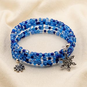 May include: A multi-strand wrap bracelet featuring various shades of blue and clear glass beads. The bracelet includes two silver-toned metal snowflake charms attached to the ends, creating a layered appearance with a circular, flexible design.