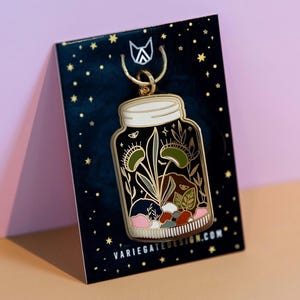 Pode incluir: Um alfinete de esmalte dourado em forma de jarra com um design de terrário com armadilhas de Vénus, plantas e pedras. O alfinete está em um cartão azul escuro com estrelas douradas e o texto "VARIEGATEDESIGN.COM".