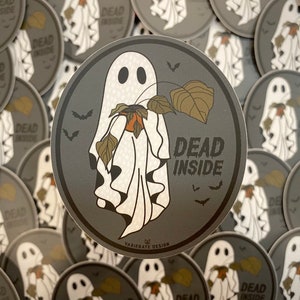 May include: A round sticker with a gray background features a white ghost holding a brown leaf. The ghost has a white sheet with frills and a black face with two black eyes. The text "DEAD INSIDE" is written in white letters on the sticker.