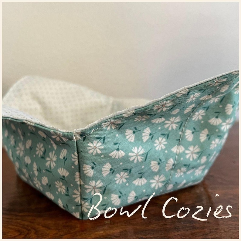 May include: A square bowl cozy with a teal floral pattern. The exterior features white flowers and green leaves on a teal background. The interior is a light cream color with a small white dot pattern. The words "Bowl Cozies" are written in white script.