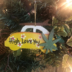 May include: A yellow camper van Christmas ornament with the text "High Love You" and a green glittery cannabis leaf. The ornament is decorated with small white and red flowers and a brown roof rack.