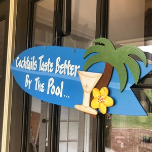 Op de afbeelding: Een blauw en wit surfboard-vormig bord met de tekst "Cocktails Taste Better By The Pool..." en een gele bloem en een martini-glas illustratie.