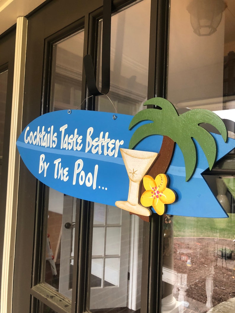 Op de afbeelding: Een blauw en wit surfboard-vormig bord met de tekst "Cocktails Taste Better By The Pool..." en een gele bloem en een martini-glas illustratie.