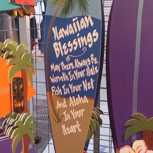 May include: A blue and brown surfboard-shaped wooden sign with the text "Hawaiian Blessings" and a palm tree design. The sign has a message that reads "May There Always Be Warmth In Your Hale, Fish In Your Net And Aloha In Your Heart."