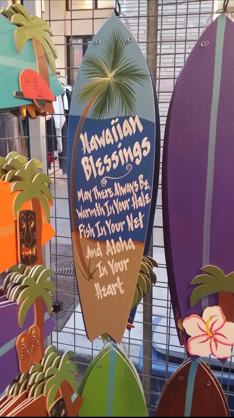 May include: A blue and brown surfboard-shaped wooden sign with the text "Hawaiian Blessings" and a palm tree design. The sign has a message that reads "May There Always Be Warmth In Your Hale, Fish In Your Net And Aloha In Your Heart."