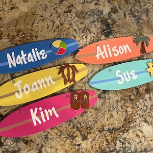 May include: Five wooden surfboard-shaped signs with different names and beach-themed decorations. The signs are painted in various colors, including blue, yellow, pink, orange, and turquoise. The decorations include a beach ball, a turtle, flip-flops, a palm tree, and a sun.