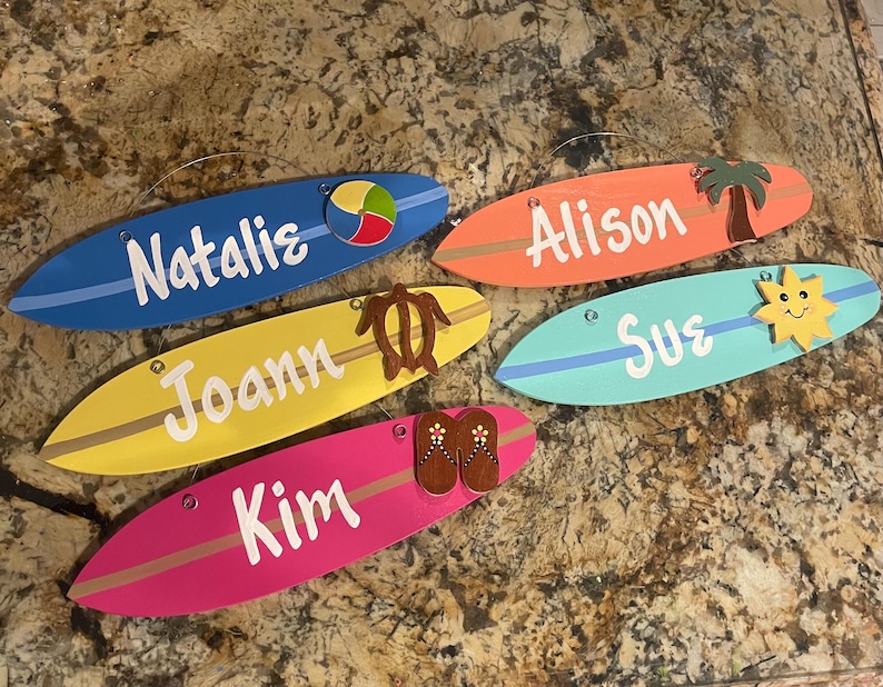 May include: Five wooden surfboard-shaped signs with different names and beach-themed decorations. The signs are painted in various colors, including blue, yellow, pink, orange, and turquoise. The decorations include a beach ball, a turtle, flip-flops, a palm tree, and a sun.
