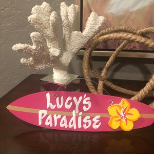 May include: Pink surfboard-shaped sign with the text "Lucy's Paradise" in white paint. A yellow hibiscus flower is attached to the right side of the sign.