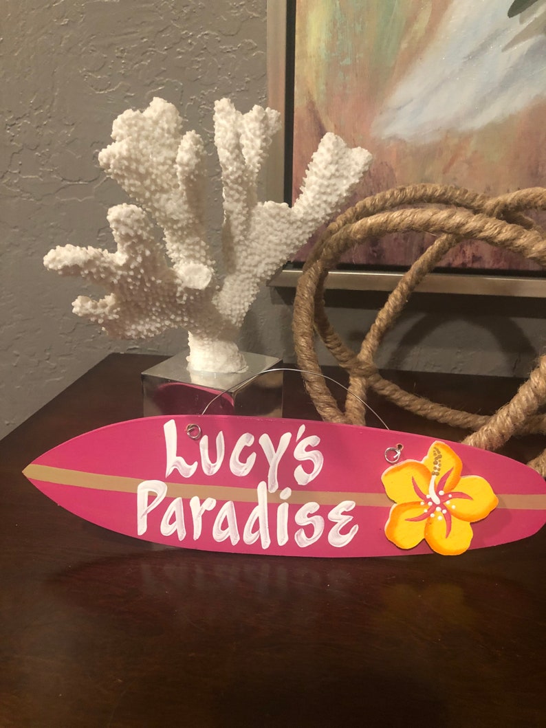 May include: Pink surfboard-shaped sign with the text "Lucy's Paradise" in white paint. A yellow hibiscus flower is attached to the right side of the sign.