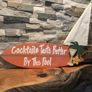 Op de afbeelding: Een houten surfboard-bord met de tekst "Cocktails Taste Better By The Pool..." in wit geschilderd. Het bord is oranje geverfd en heeft een bruine streep. Een kleine gele bloem en een palmboom zijn op het bord geschilderd.