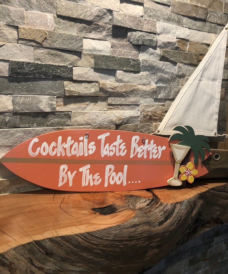 Op de afbeelding: Een houten surfboard-bord met de tekst "Cocktails Taste Better By The Pool..." in wit geschilderd. Het bord is oranje geverfd en heeft een bruine streep. Een kleine gele bloem en een palmboom zijn op het bord geschilderd.