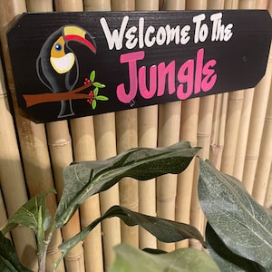 May include: A black wooden sign with the text "Welcome to the Jungle" in white and pink lettering. A toucan with a yellow beak and black and white feathers is perched on a branch with red berries.
