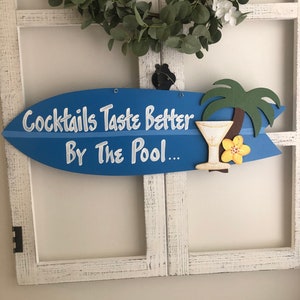 Op de afbeelding: Blauw surfplank-vormig houten bord met de tekst "Cocktails Taste Better By The Pool..." in witte verf. Een gele bloem en een palmboom zijn op het bord geschilderd, samen met een blauw en wit cocktailglas.