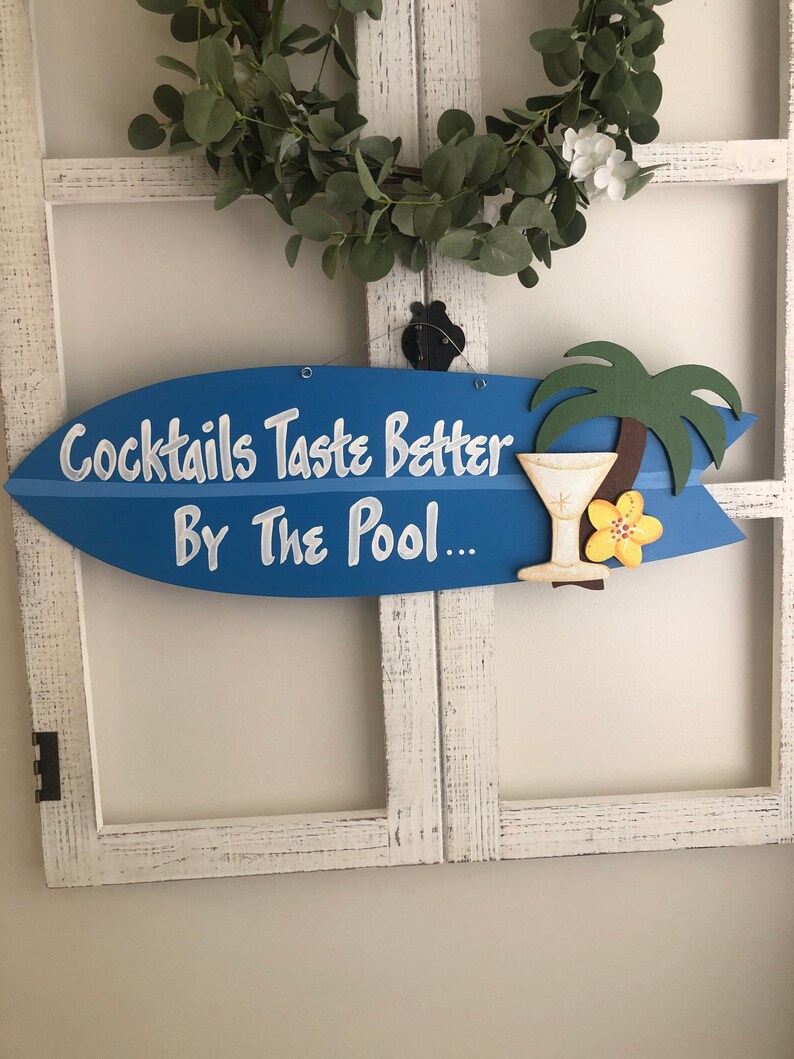 Op de afbeelding: Blauw surfplank-vormig houten bord met de tekst "Cocktails Taste Better By The Pool..." in witte verf. Een gele bloem en een palmboom zijn op het bord geschilderd, samen met een blauw en wit cocktailglas.