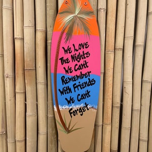 May include: A wooden surfboard-shaped sign with a painted palm tree, ocean waves, and the text "We love the nights we can't remember with friends we can't forget."