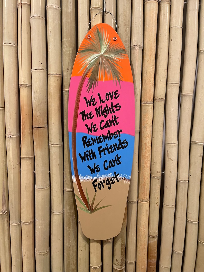 May include: A wooden surfboard-shaped sign with a painted palm tree, ocean waves, and the text "We love the nights we can't remember with friends we can't forget."