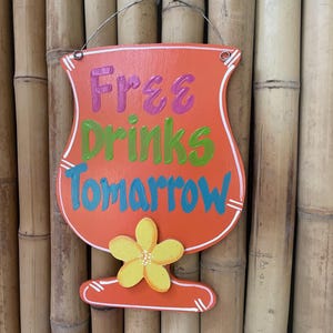 May include: Orange wooden sign shaped like a cocktail glass with the text "Free Drinks Tomorrow" in pink, green, and blue lettering. A yellow flower is painted on the bottom of the glass.