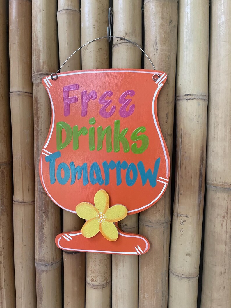 May include: Orange wooden sign shaped like a cocktail glass with the text "Free Drinks Tomorrow" in pink, green, and blue lettering. A yellow flower is painted on the bottom of the glass.