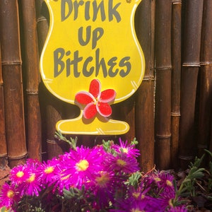 May include: A yellow wooden sign shaped like a cocktail glass with the text "Drink Up Bitches" in black lettering. A red hibiscus flower is painted on the sign. The sign is hanging on a bamboo fence with purple flowers in the foreground.