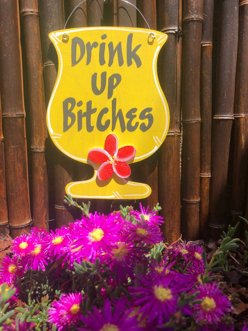 May include: A yellow wooden sign shaped like a cocktail glass with the text "Drink Up Bitches" in black lettering. A red hibiscus flower is painted on the sign. The sign is hanging on a bamboo fence with purple flowers in the foreground.