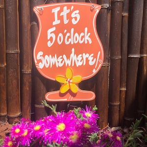 May include: A wooden sign shaped like a cocktail glass with the text "It's 5 o'clock Somewhere!" in white paint. The sign is hanging on a bamboo fence with pink flowers in the foreground.