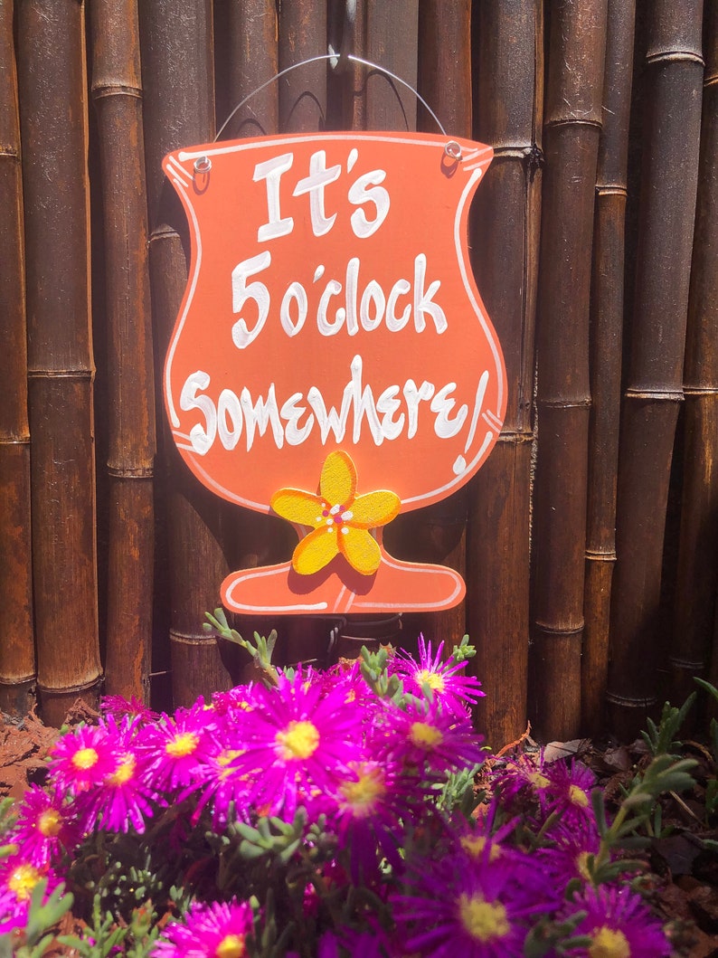 May include: A wooden sign shaped like a cocktail glass with the text "It's 5 o'clock Somewhere!" in white paint. The sign is hanging on a bamboo fence with pink flowers in the foreground.