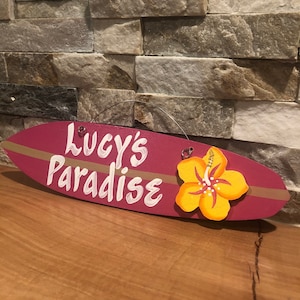 May include: A pink surfboard-shaped wooden sign with the text "Lucy's Paradise" in white paint. A yellow hibiscus flower is attached to the sign.