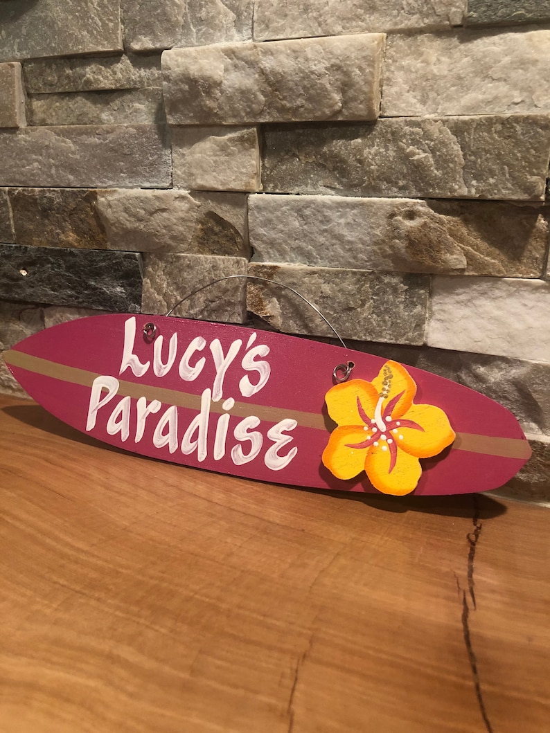 May include: A pink surfboard-shaped wooden sign with the text "Lucy's Paradise" in white paint. A yellow hibiscus flower is attached to the sign.