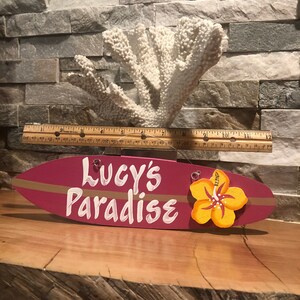 May include: A pink surfboard-shaped wooden sign with the text "Lucy's Paradise" in white paint and a yellow hibiscus flower painted on the right side.
